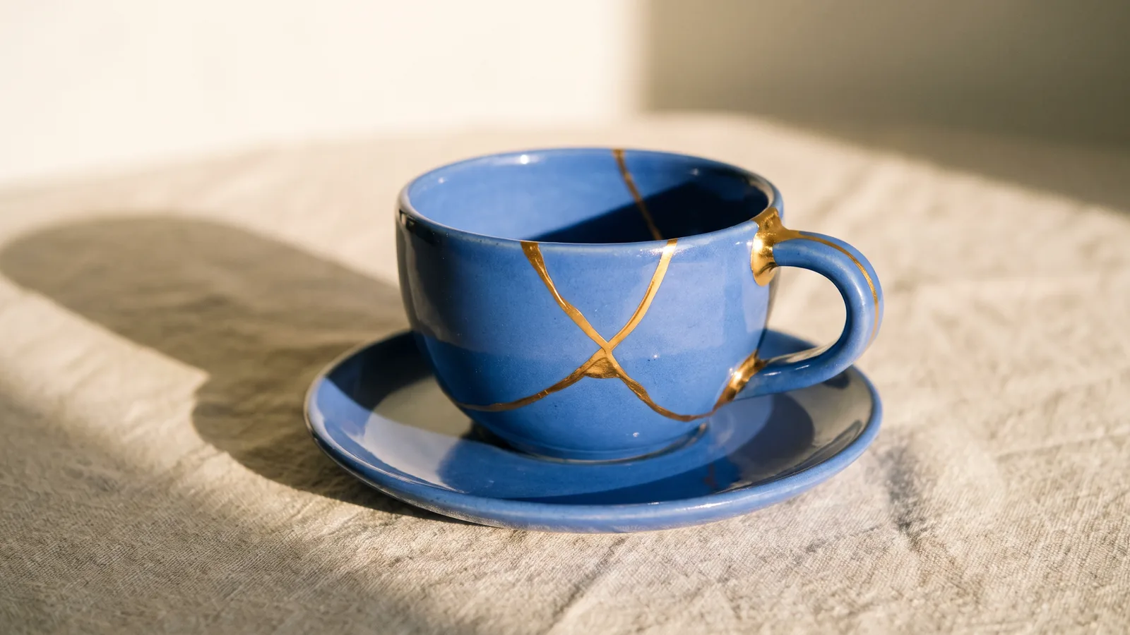 A cornflower blue cup and saucer, its cracks mended with gold, sits on a linen tablecloth in the morning sun.