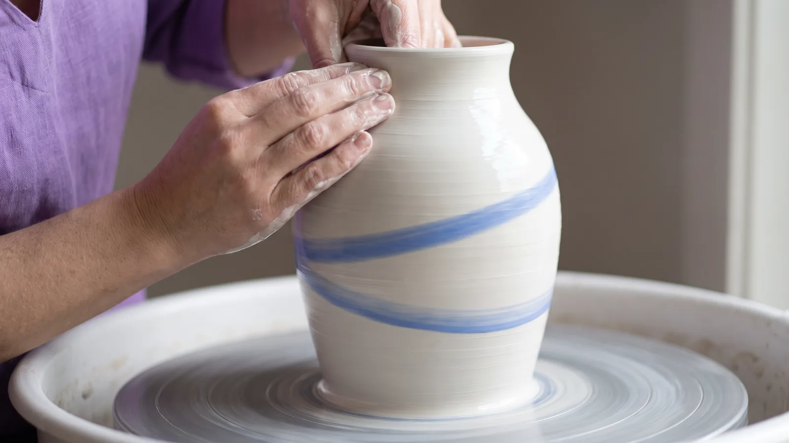 Wet hands shape a white clay vase with a blue spiral band on a spinning potter's wheel.