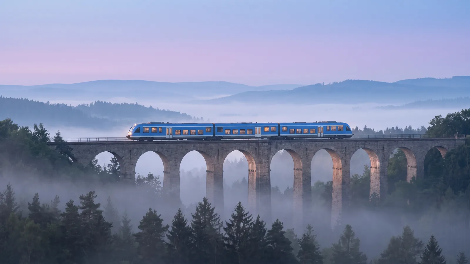 A blue regional train crosses a stone arch viaduct above a forested valley in the morning mist.