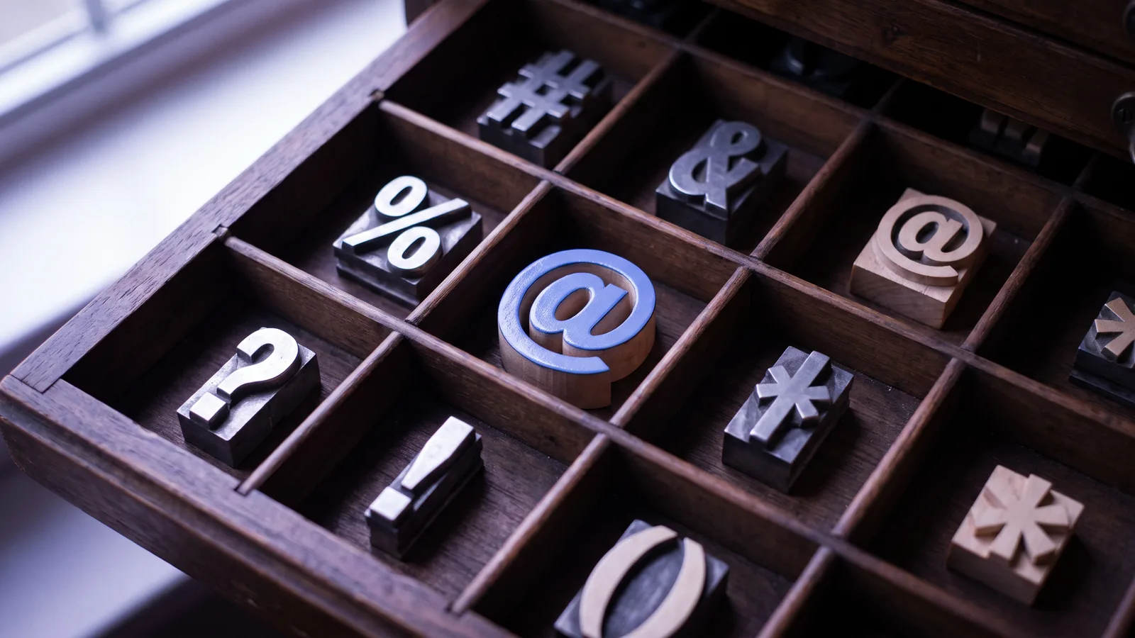 An antique printer's type case filled with symbol type: a hash, an ampersand, a percent sign, question and exclamation marks, and a blue at sign in the middle.
