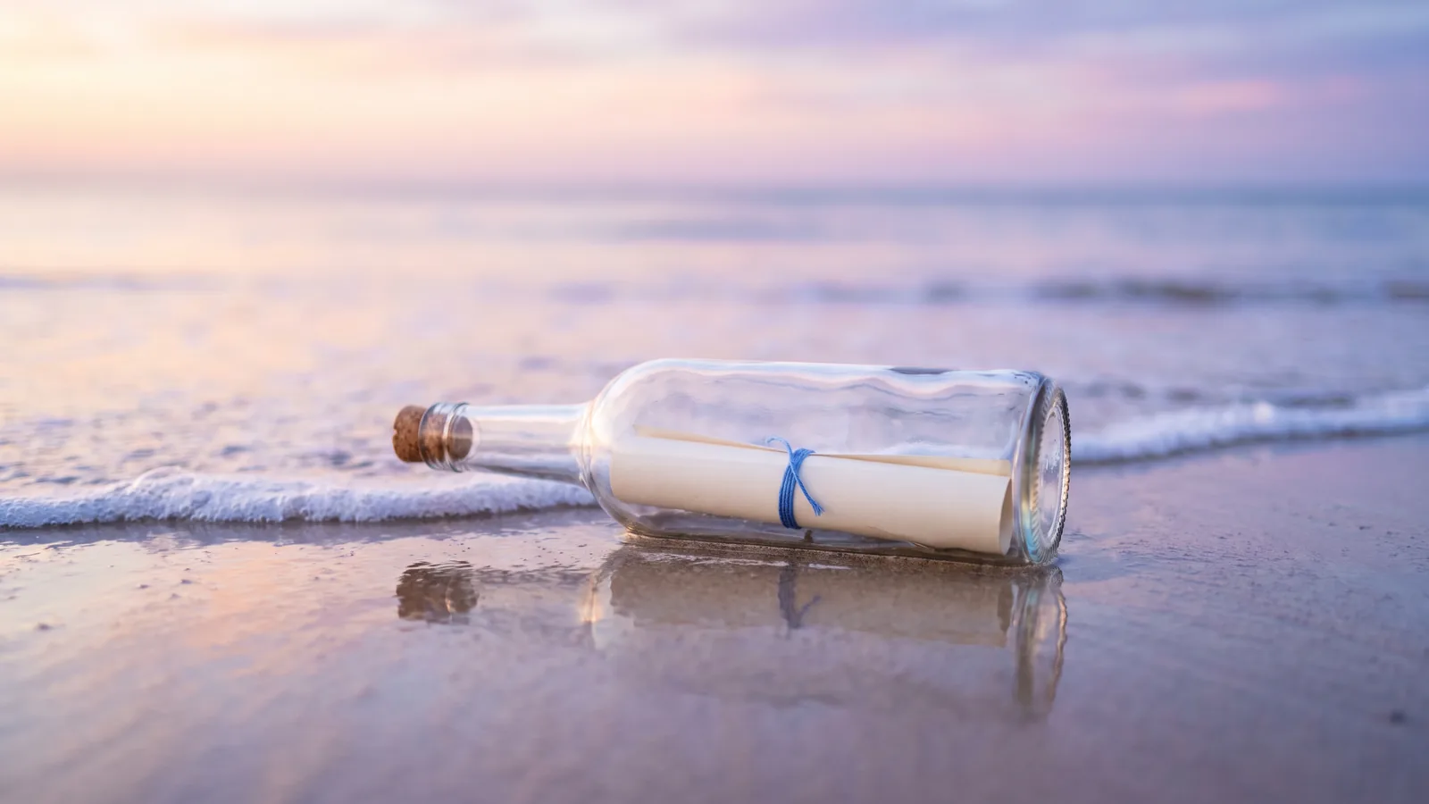 A corked glass bottle holding a rolled message tied with blue thread lies on wet sand at the water’s edge at dawn.
