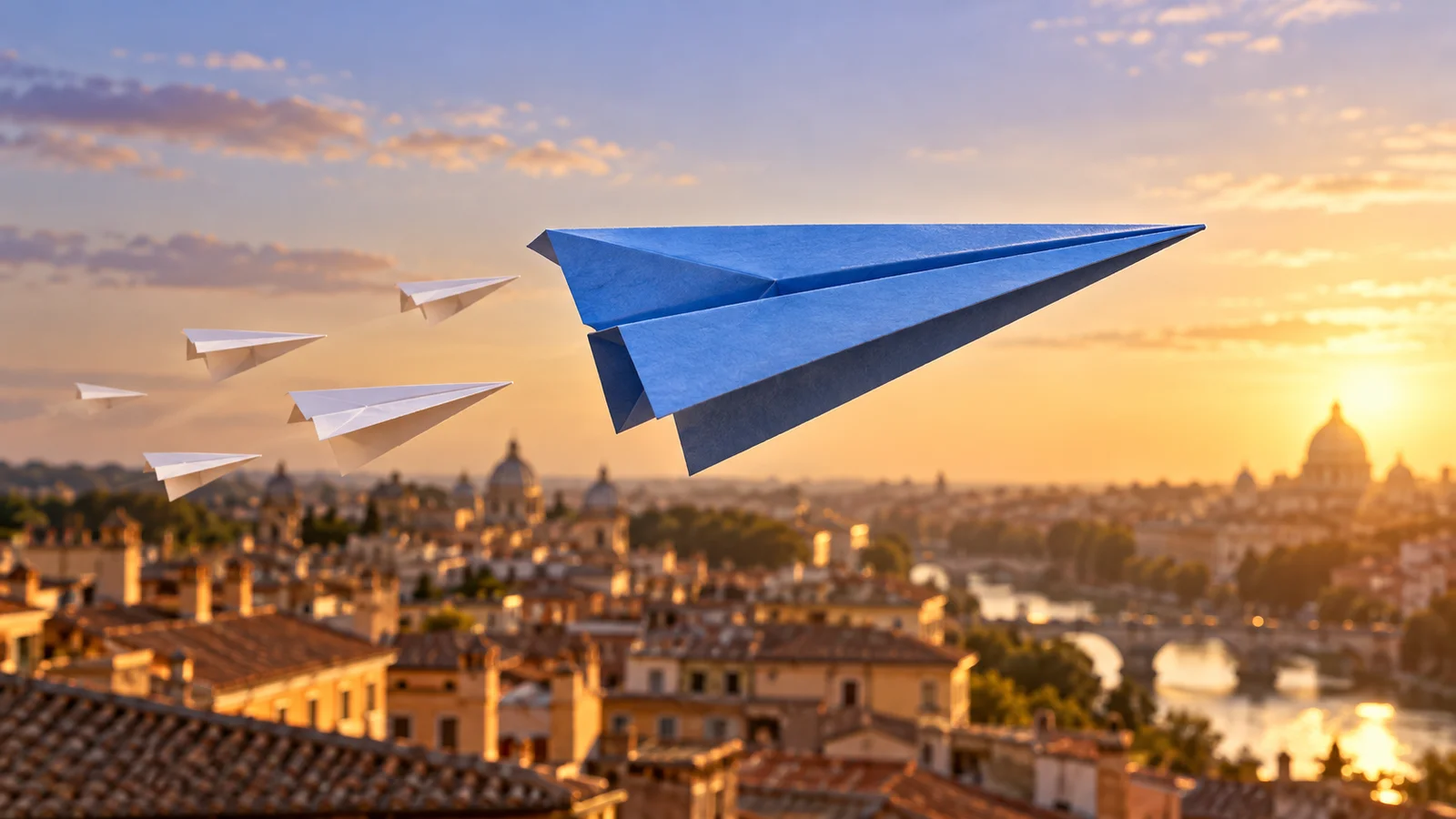 A blue paper airplane leads white paper airplanes above rooftops at sunset.